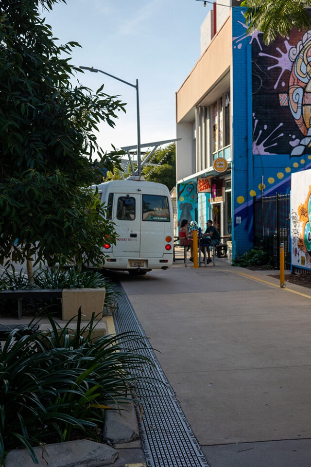 A white bus runs over drainage grates in an alleyway surrounded by greenery and mural covered walls