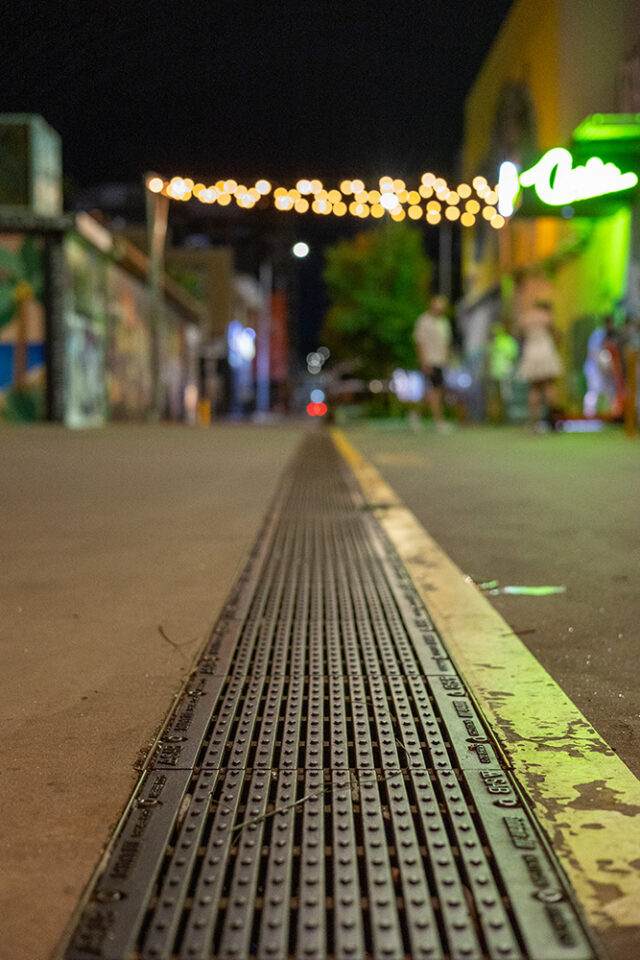 Cast iron drain grate with anti-slip nodules is photographed in a well lit alleyway in Darwin