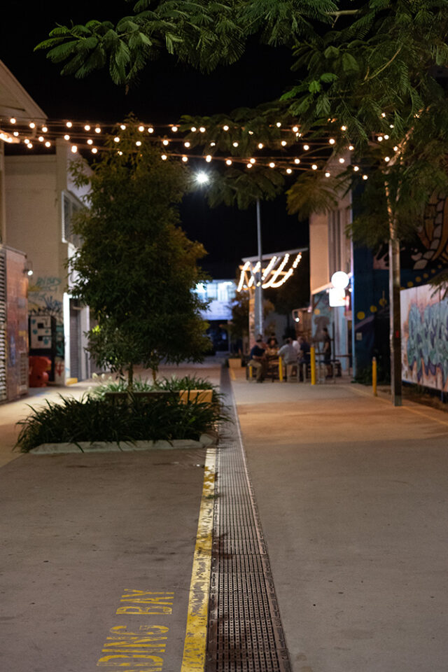 A well-lit alley at night with string lights, plants, and trees lining the path. Some people are seated in the background at a bar. Cast iron drainage grates are installed in the concrete, running the length of the alleyway