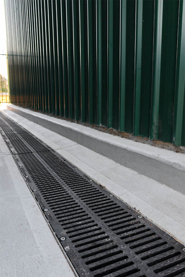 A sloped concrete surface with a cast iron drainage grate runs alongside a tall, green corrugated metal wall